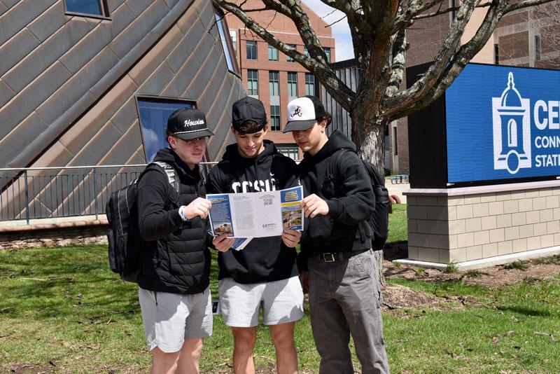 Three male students outside student center reading polytech brochure