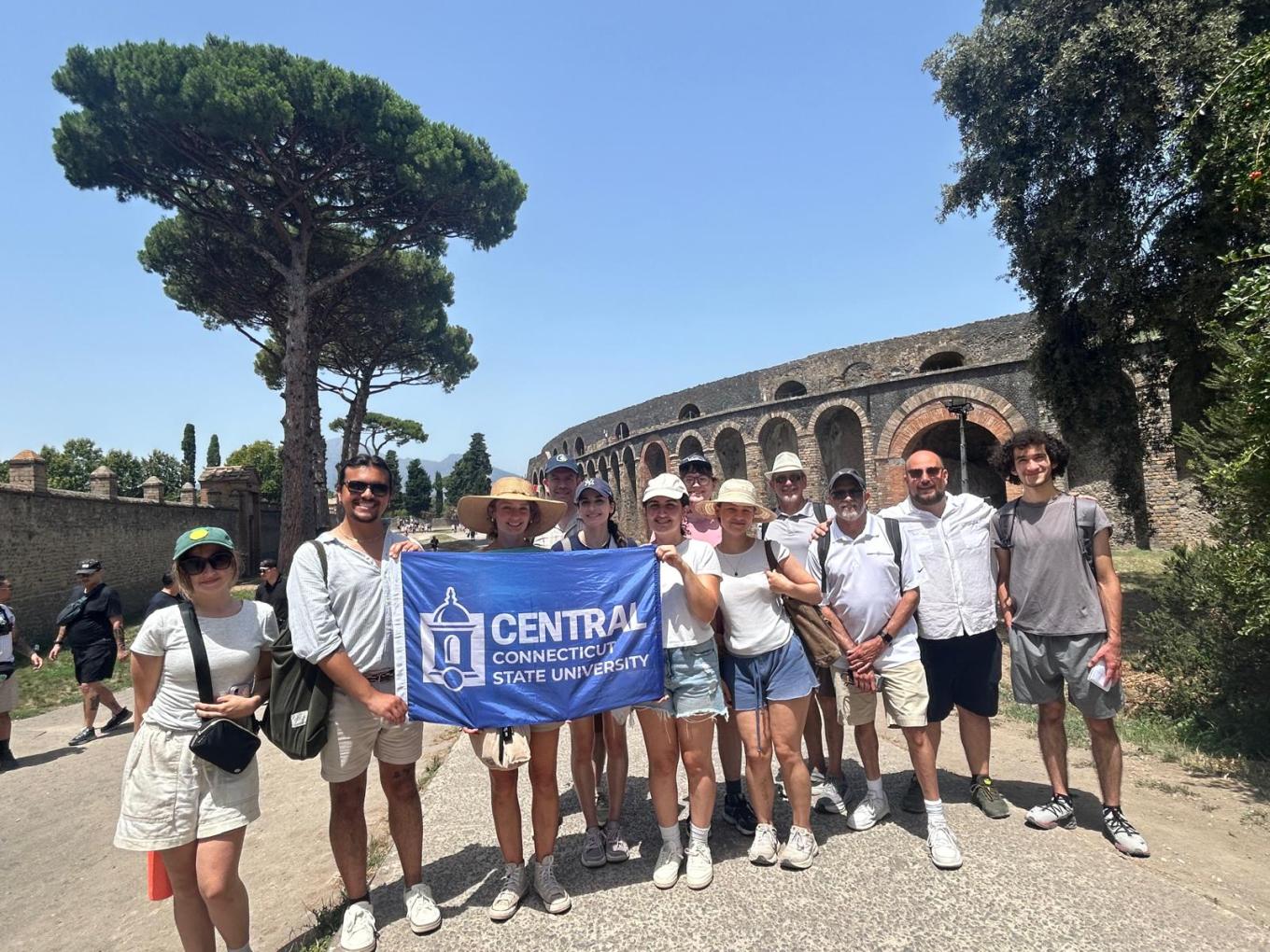 A group of Central students hold a Central banner at a historic site in the Amalfi region of Italy.