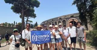 A group of Central students hold a Central banner at a historic site in the Amalfi region of Italy.